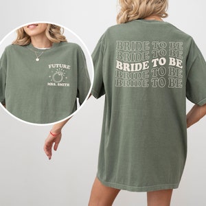 May include: Olive green t-shirt with white text. The front of the shirt says "FUTURE MRS. SMITH" with a ring graphic. The back of the shirt says "BRIDE TO BE" repeated multiple times. A casual, oversized fit.