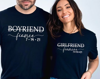 May include: Two black short-sleeved t-shirts featuring white text. The left shirt displays the word BOYFRIEND with a line through it, followed by the word fiance and the date 7-14-21. The right shirt displays GIRLFRIEND with a line through it, fiancee, and 7-14-21.