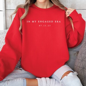 May include: Red sweatshirt with white text that reads "In my engaged era 07.11.23".