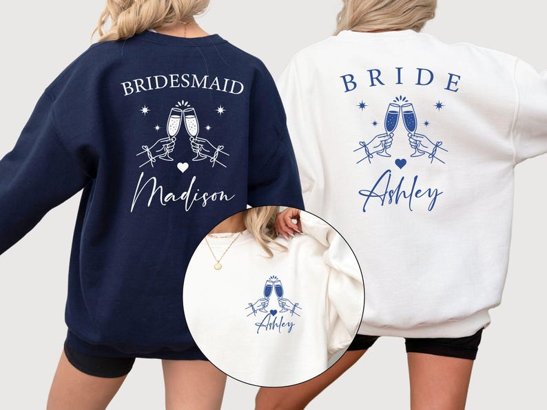 May include: Navy blue and white sweatshirts with matching designs for a bridal party. The navy sweatshirt says "Bridesmaid" with a champagne toast graphic and the name "Madison". The white sweatshirt says "Bride" with a champagne toast graphic and the name "Ashley".