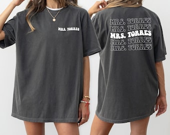 May include: A dark gray oversized t-shirt with the text "MRS. TORRES" printed on the front and back. The text is in a white retro font.