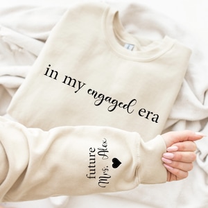 Puede incluir: Sudadera beige con el texto "in my engaged era" en escritura negra. La manga presenta el texto "future Mrs. Alex" con un s&iacute;mbolo de coraz&oacute;n negro. La sudadera est&aacute; doblada sobre una superficie blanca.