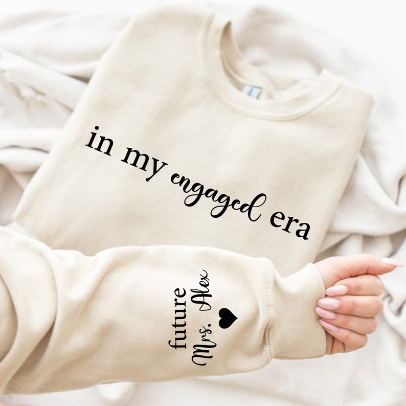 Puede incluir: Sudadera beige con el texto "in my engaged era" en escritura negra. La manga presenta el texto "future Mrs. Alex" con un s&iacute;mbolo de coraz&oacute;n negro. La sudadera est&aacute; doblada sobre una superficie blanca.