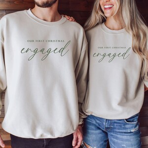 May include: Two beige crewneck sweatshirts with the text "OUR FIRST CHRISTMAS" above the word "engaged" in green script. The sweatshirts are worn by two people, one wearing a bracelet. The background is a wooden wall.