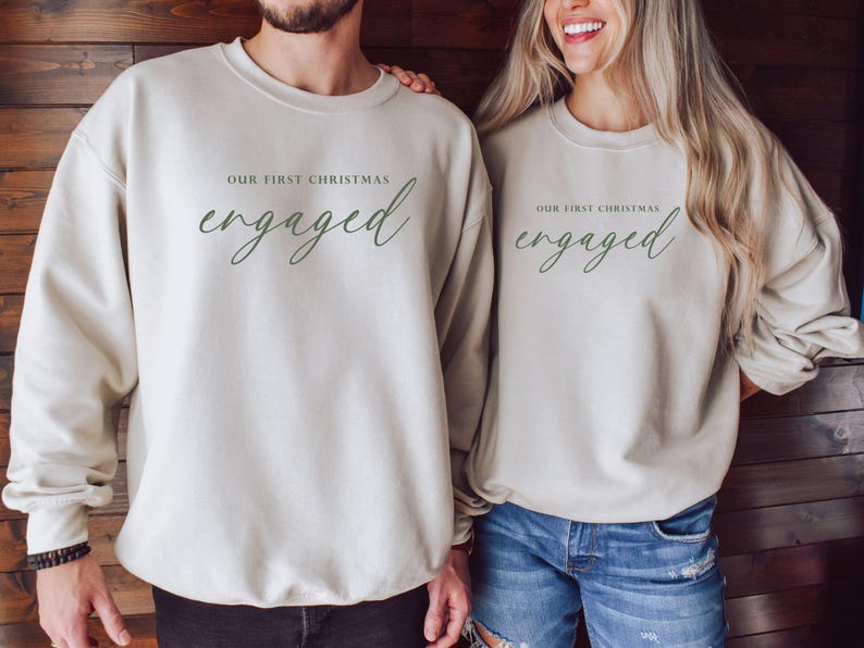 May include: Two beige crewneck sweatshirts with the text "OUR FIRST CHRISTMAS" above the word "engaged" in green script. The sweatshirts are worn by two people, one wearing a bracelet. The background is a wooden wall.