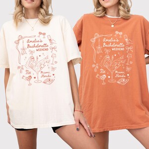 Könnte beinhalten: Zwei kurzärmelige T-Shirts mit Rundhalsausschnitt in Creme und Rostorange, bedruckt mit einer roten Strichgrafik. Das Design zeigt den Text "Amelia's Bachelorette Weekend", "dec 2026" und "Miami, Florida" sowie Abbildungen eines Martiniglases, Schleifen und einer Braut.