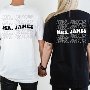 K&ouml;nnte beinhalten: Zwei schwarz-wei&szlig;e T-Shirts mit dem Text "MR. JAMES" und "MRS. JAMES", der jeweils f&uuml;nfmal auf jedem Shirt wiederholt wird.