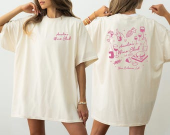 May include: A white oversized t-shirt with a pink graphic design on the back. The design features wine glasses, bottles, grapes, and cheese. The text on the shirt reads "Amelia's Wine Club est. 2023 New Orleans, LA."