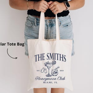 May include: Cream-colored tote bag with navy blue text and illustrations. The bag features the words "THE SMITHS" above a cocktail and other graphics, with "Honeymoon Club" and "MIAMI, FL" below. The year "2024" is also printed on the bag.
