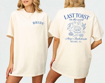 May include: Two white t-shirts with blue text and graphics. The front shirt says "Bride". The back shirt says "Last Toast on the coast" with a graphic of seashells, starfish, and drinks. The text below the graphic says "Amy's Bachelorette Miami, FL."