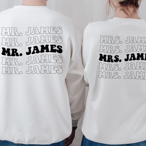 K&ouml;nnte beinhalten: Zwei wei&szlig;e Sweatshirts mit schwarzem Text. Der Text auf dem linken Sweatshirt lautet "MR. JAMES" f&uuml;nfmal. Der Text auf dem rechten Sweatshirt lautet "MRS. JAMES" f&uuml;nfmal.