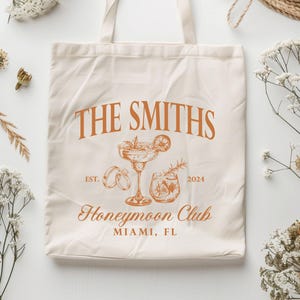May include: Cream-colored tote bag with the text "THE SMITHS" in orange, along with illustrations of a cocktail, rings, and a drink. The text "Honeymoon Club" and "MIAMI, FL" are also printed on the bag. The year "2024" is included.
