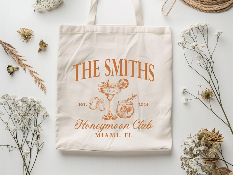 May include: Cream-colored tote bag with the text "THE SMITHS" in orange, along with illustrations of a cocktail, rings, and a drink. The text "Honeymoon Club" and "MIAMI, FL" are also printed on the bag. The year "2024" is included.