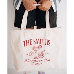 May include: Cream-colored tote bag with a red graphic design. The design includes the text "THE SMITHS", "Honeymoon Club", and "Miami, FL", along with illustrations of cocktails and rings. The year "2024" is also included.