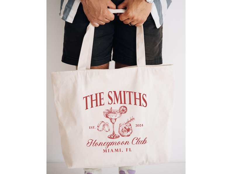 May include: Cream-colored tote bag with a red graphic design. The design includes the text "THE SMITHS", "Honeymoon Club", and "Miami, FL", along with illustrations of cocktails and rings. The year "2024" is also included.