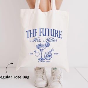 K&ouml;nnte beinhalten: Cremefarbene Tragetasche mit blauem Text und Illustrationen. Der Text lautet "THE FUTURE Mrs. Miller" mit Cocktail, Ring und anderen Grafiken. Der Text unten lautet "I SAID YES!" und "2024".