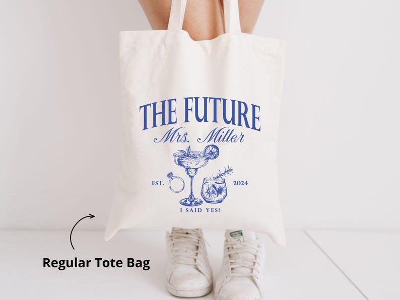 K&ouml;nnte beinhalten: Cremefarbene Tragetasche mit blauem Text und Illustrationen. Der Text lautet "THE FUTURE Mrs. Miller" mit Cocktail, Ring und anderen Grafiken. Der Text unten lautet "I SAID YES!" und "2024".