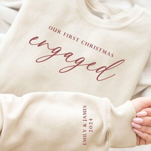 May include: Cream-colored sweatshirt with the text "OUR FIRST CHRISTMAS engaged" in burgundy script. The sleeve displays "EMILY & JAMES 2024" in burgundy. A cozy, casual top for celebrating an engagement.