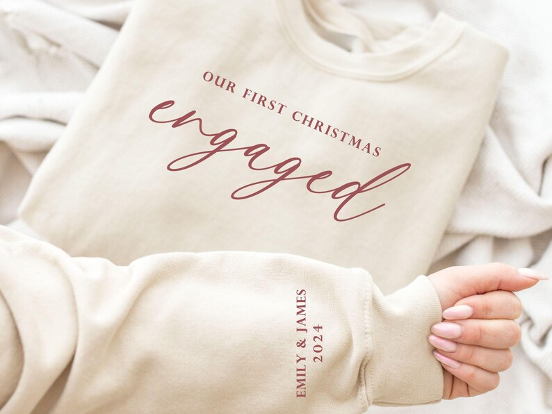 May include: Cream-colored sweatshirt with the text "OUR FIRST CHRISTMAS engaged" in burgundy script. The sleeve displays "EMILY & JAMES 2024" in burgundy. A cozy, casual top for celebrating an engagement.