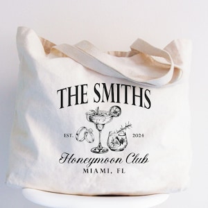 May include: Cream-colored tote bag with the text "THE SMITHS" above a cocktail glass, rings, and a drink. Below, it reads "Honeymoon Club" and "MIAMI, FL". The bag has a simple design with a handle.