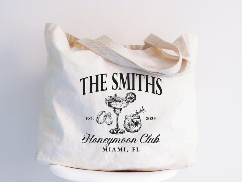 May include: Cream-colored tote bag with the text "THE SMITHS" above a cocktail glass, rings, and a drink. Below, it reads "Honeymoon Club" and "MIAMI, FL". The bag has a simple design with a handle.