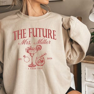 May include: Beige sweatshirt with the text "The Future Mrs. Miller" in red, with a ring, a margarita glass, and a cocktail glass printed on the front. The text "Est. 2024" and "I said yes!" are also printed on the sweatshirt.