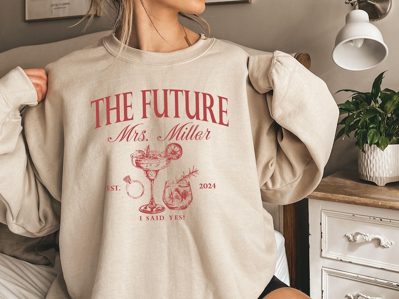 May include: Beige sweatshirt with the text "The Future Mrs. Miller" in red, with a ring, a margarita glass, and a cocktail glass printed on the front. The text "Est. 2024" and "I said yes!" are also printed on the sweatshirt.