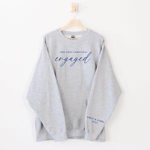 May include: A gray sweatshirt with the words "OUR FIRST CHRISTMAS" above the word "engaged" in navy blue script. The sleeve has "EMILY & JAMES 2024" printed in navy blue. The sweatshirt is hanging on a wooden hanger.