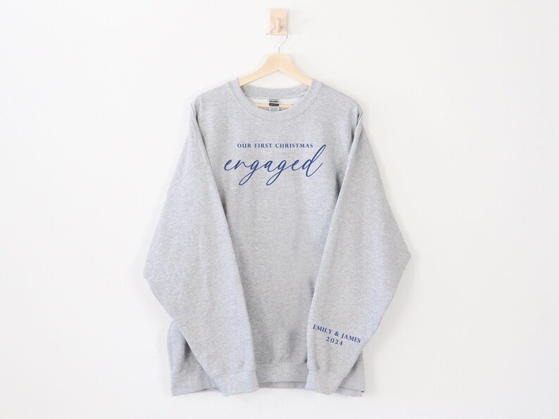 May include: A gray sweatshirt with the words "OUR FIRST CHRISTMAS" above the word "engaged" in navy blue script. The sleeve has "EMILY & JAMES 2024" printed in navy blue. The sweatshirt is hanging on a wooden hanger.