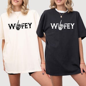 May include: Two short-sleeved t-shirts, one cream and one black, each with the word "Wifey" in bold white letters and a skeleton hand gesture. The shirts are a casual style, suitable for everyday wear.