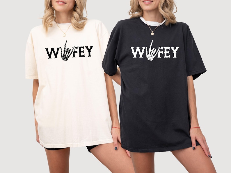 May include: Two short-sleeved t-shirts, one cream and one black, each with the word "Wifey" in bold white letters and a skeleton hand gesture. The shirts are a casual style, suitable for everyday wear.