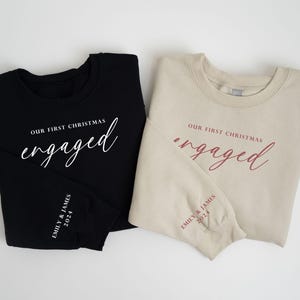 May include: Two sweatshirts, one black and one beige, with the words "Our First Christmas engaged" printed on the front in white and burgundy script. The sleeves have "Emily & James 2024" printed on them. A festive holiday theme.