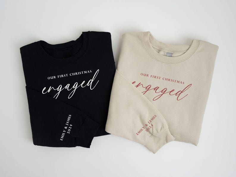 May include: Two sweatshirts, one black and one beige, with the words "Our First Christmas engaged" printed on the front in white and burgundy script. The sleeves have "Emily & James 2024" printed on them. A festive holiday theme.