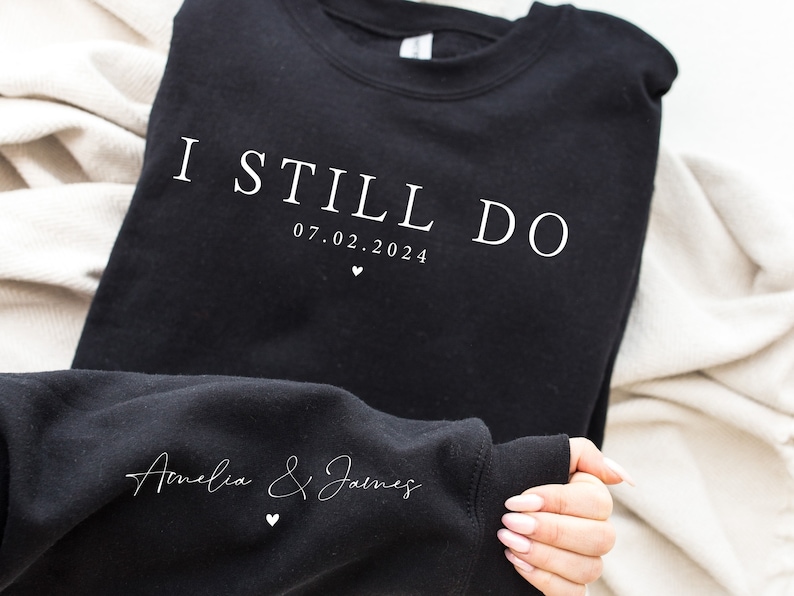 May include: Black sweatshirt with white text that reads "I Still Do 07.02.2024" and "Amelia & James" with a small heart symbol.