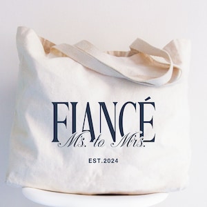 May include: Cream-colored canvas tote bag with the words "FIANCÉ Ms. to Mrs. EST.2024" printed in navy blue. The bag has matching cream-colored handles and is likely used for carrying items. A wedding-themed gift.