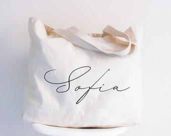 May include: A cream-colored canvas tote bag featuring two fabric handles and the name Sofia printed in a black cursive script across the center of the front panel. The bag has a soft, unstructured shape and a simple, clean design.