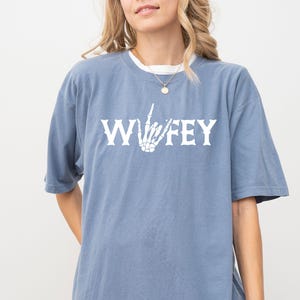 May include: A dusty blue t-shirt with the word "Wifey" in white block letters and a white skeleton hand making a gesture. The shirt has short sleeves and a relaxed fit.