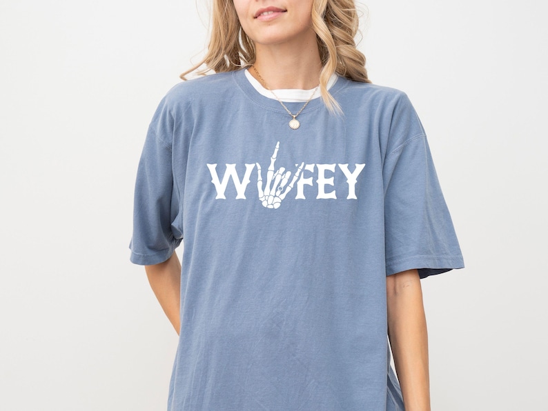May include: A dusty blue t-shirt with the word "Wifey" in white block letters and a white skeleton hand making a gesture. The shirt has short sleeves and a relaxed fit.