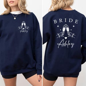 May include: Navy blue sweatshirt with white graphic. Front features two champagne glasses clinking, hands, a heart, and the name "Ashley". The back says "BRIDE" above the same champagne glasses, stars, a heart, and "Ashley".