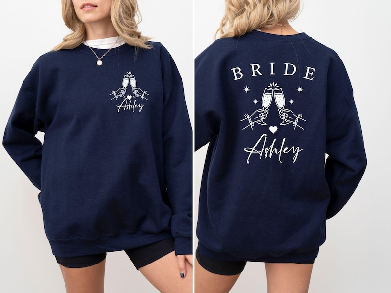 May include: Navy blue sweatshirt with white graphic. Front features two champagne glasses clinking, hands, a heart, and the name "Ashley". The back says "BRIDE" above the same champagne glasses, stars, a heart, and "Ashley".