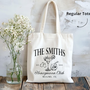 May include: Cream-colored tote bag with black text and illustrations. The text reads "THE SMITHS" above a cocktail illustration, "EST. 2024" and "Honeymoon Club MIAMI, FL". A clear glass vase with white flowers sits to the left.