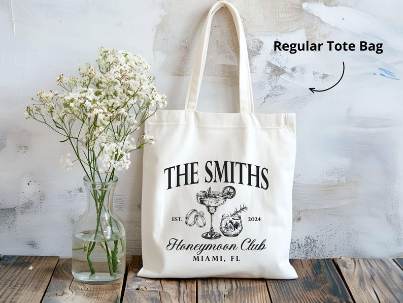 May include: Cream-colored tote bag with black text and illustrations. The text reads "THE SMITHS" above a cocktail illustration, "EST. 2024" and "Honeymoon Club MIAMI, FL". A clear glass vase with white flowers sits to the left.
