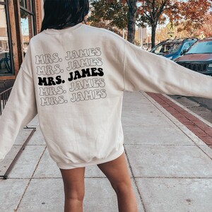 K&ouml;nnte beinhalten: Ein wei&szlig;er Pullover mit dem Text "MRS. JAMES" f&uuml;nfmal in einer Retro-Schrift auf der R&uuml;ckseite.