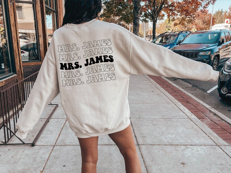 K&ouml;nnte beinhalten: Ein wei&szlig;er Pullover mit dem Text "MRS. JAMES" f&uuml;nfmal in einer Retro-Schrift auf der R&uuml;ckseite.