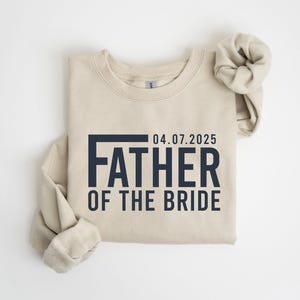 May include: A beige sweatshirt with the text "04.07.2025 FATHER OF THE BRIDE" in navy blue. The sleeves are rolled up, and the garment is laid flat on a white surface. The sweatshirt is a casual garment.