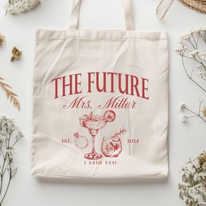 K&ouml;nnte beinhalten: Cremefarbene Tragetasche mit rotem Text und Illustrationen. Der Text lautet "THE FUTURE Mrs. Miller" mit "EST. 2024 I SAID YES!" unter einer Cocktail- und Ring-Illustration. Die Tasche hat zwei Griffe.