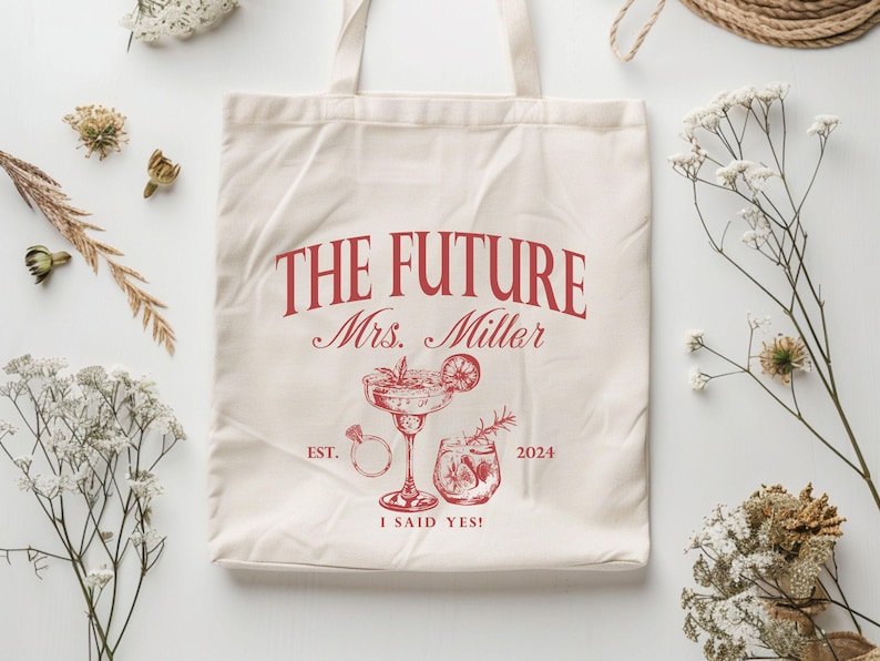 K&ouml;nnte beinhalten: Cremefarbene Tragetasche mit rotem Text und Illustrationen. Der Text lautet "THE FUTURE Mrs. Miller" mit "EST. 2024 I SAID YES!" unter einer Cocktail- und Ring-Illustration. Die Tasche hat zwei Griffe.