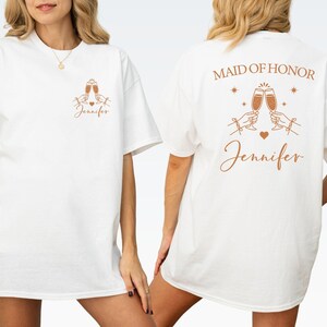 May include: White t-shirt with a front and back design. The front features two champagne glasses, a heart, and the name "Jennifer" in a script font. The back says "Maid of Honor" above the same champagne glasses and name.