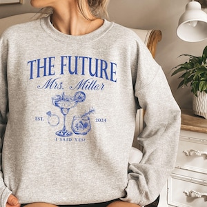 May include: Gray sweatshirt with blue text that reads "The Future Mrs. Miller Est. 2024 I Said Yes!" with a drawing of a cocktail glass and a ring.