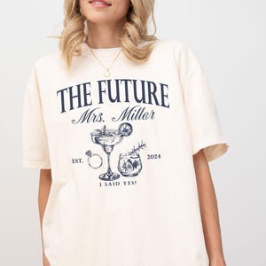May include: A white t-shirt with the text "The Future Mrs. Miller Est. 2024 I Said Yes!" printed on it. The design also includes a cocktail glass with a ring and a small glass of liquid.